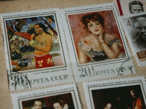 A collection of vintage postage stamps featuring artworks and portraits. One stamp displays a person in a vibrant red garment, surrounded by natural scenery. Another features a woman with red hair, adorned in an elegant dress, posing thoughtfully. Additional stamps feature monochromatic portraits of individuals from history.