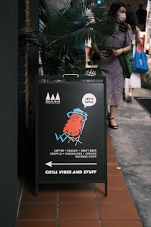 A sidewalk sign with an illustration of a relaxed character lounging in a chair. The sign advertises a place offering coffee, coolers, craft beer, croffle, cheesecake, cookies, and outdoor items. The slogan 'CHILL VIBES AND STUFF' is prominently displayed. A person wearing a mask and holding a shopping bag is walking past the sign.