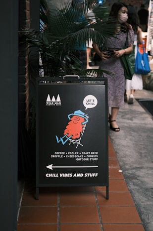 A sidewalk sign with an illustration of a relaxed character lounging in a chair. The sign advertises a place offering coffee, coolers, craft beer, croffle, cheesecake, cookies, and outdoor items. The slogan 'CHILL VIBES AND STUFF' is prominently displayed. A person wearing a mask and holding a shopping bag is walking past the sign.