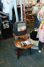A vintage television is displaying a close-up of a woman's face. The TV is placed on a wooden round table, which also holds a collection of VHS tapes. In the background, there are racks of vintage clothing, including a sports jersey and various shirts. A person with a tote bag is visible, browsing through items in the store.