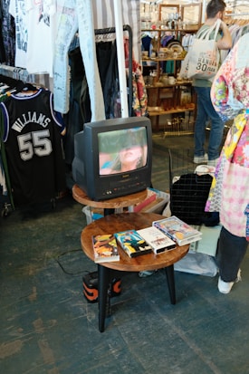 A vintage television is displaying a close-up of a woman's face. The TV is placed on a wooden round table, which also holds a collection of VHS tapes. In the background, there are racks of vintage clothing, including a sports jersey and various shirts. A person with a tote bag is visible, browsing through items in the store.