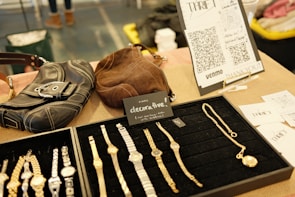 A display of wristwatches and jewelry is neatly arranged on a black velvet tray. Nearby, there are two leather bags, one black and one brown. A small sign reads 'decorative,' indicating the items are for sale, priced at $5 each. In the background, there is a stand with QR codes and the word 'thrift,' likely for digital payment options such as Venmo. Additional promotional material and business cards are visible.