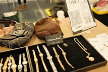 A display of wristwatches and jewelry is neatly arranged on a black velvet tray. Nearby, there are two leather bags, one black and one brown. A small sign reads 'decorative,' indicating the items are for sale, priced at $5 each. In the background, there is a stand with QR codes and the word 'thrift,' likely for digital payment options such as Venmo. Additional promotional material and business cards are visible.