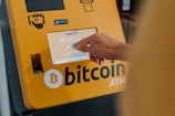 A hand interacts with a touchscreen on a Bitcoin ATM, which is yellow and prominently displays a Bitcoin logo. The screen shows options related to purchasing cryptocurrency.