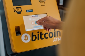 A hand interacts with a touchscreen on a Bitcoin ATM, which is yellow and prominently displays a Bitcoin logo. The screen shows options related to purchasing cryptocurrency.