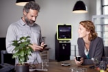 A man and a woman are interacting in a modern, well-lit space. The man is looking down at a device in his hands, possibly a smartphone, while the woman is smiling at him, holding a small cup. The background includes hanging lights, a potted plant, and a device resembling an ATM or kiosk.