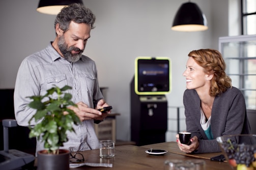 A man and a woman are interacting in a modern, well-lit space. The man is looking down at a device in his hands, possibly a smartphone, while the woman is smiling at him, holding a small cup. The background includes hanging lights, a potted plant, and a device resembling an ATM or kiosk.