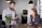 A man and a woman are interacting in a modern, well-lit space. The man is looking down at a device in his hands, possibly a smartphone, while the woman is smiling at him, holding a small cup. The background includes hanging lights, a potted plant, and a device resembling an ATM or kiosk.
