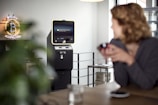 A cryptocurrency ATM is situated in a modern indoor setting. A neon sign that reads 'Bitcoin Accepted Here' is prominently displayed on the wall. A person in the foreground appears slightly blurred, sitting at a table with a cup. The room has a bright and minimalistic design with a clean, industrial look.