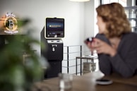A cryptocurrency ATM is situated in a modern indoor setting. A neon sign that reads 'Bitcoin Accepted Here' is prominently displayed on the wall. A person in the foreground appears slightly blurred, sitting at a table with a cup. The room has a bright and minimalistic design with a clean, industrial look.