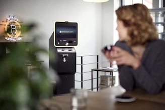 A cryptocurrency ATM is situated in a modern indoor setting. A neon sign that reads 'Bitcoin Accepted Here' is prominently displayed on the wall. A person in the foreground appears slightly blurred, sitting at a table with a cup. The room has a bright and minimalistic design with a clean, industrial look.