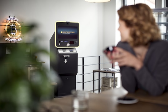 A cryptocurrency ATM is situated in a modern indoor setting. A neon sign that reads 'Bitcoin Accepted Here' is prominently displayed on the wall. A person in the foreground appears slightly blurred, sitting at a table with a cup. The room has a bright and minimalistic design with a clean, industrial look.