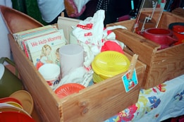 A wooden box containing a collection of vintage items, including a children's book titled 'We Help Mommy,' stacks of plates, a plastic cup, and a few colorful lids. The box sits on a table covered with a floral-patterned cloth, with other boxes and kitchenware visible in the background.