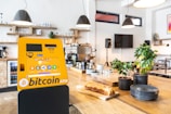 A bitcoin ATM in a modern cafe setting, positioned on a wooden table with shelves in the background. The ATM is yellow with multiple cryptocurrency logos displayed on its screen. Various potted plants and neatly stacked plates are on the table, alongside a loaf of bread on a cutting board. Overhead lights provide ambient lighting.