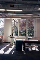 A well-lit workshop with large windows showing an outdoor view. The room contains various equipment including a drill press and a workbench with stools. Sunlight streams in, creating shadows on the floor.