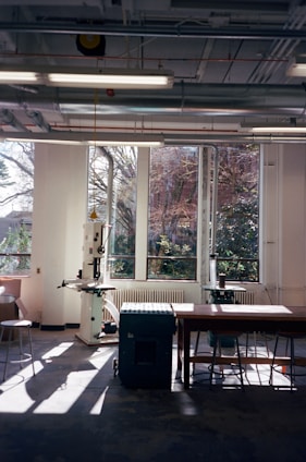 A well-lit workshop with large windows showing an outdoor view. The room contains various equipment including a drill press and a workbench with stools. Sunlight streams in, creating shadows on the floor.