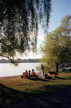 A group of people is gathered near a lakeside, sitting on portable chairs on a grassy area. A couple of people are standing near a dog. The scene is set under large overhanging tree branches, with a view of the water and distant trees in the background.