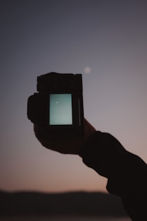 A hand is holding a camera, with its screen displaying a night sky. The sky in the background is dark, with a small bright spot resembling a star or a planet. The silhouette of distant hills or mountains is visible at the bottom.