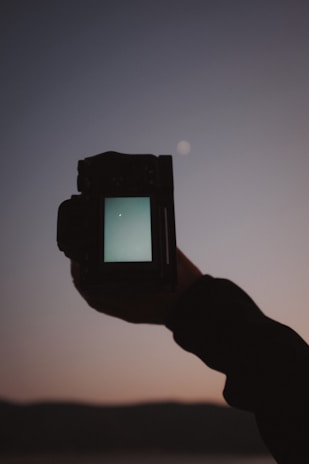 A hand is holding a camera, with its screen displaying a night sky. The sky in the background is dark, with a small bright spot resembling a star or a planet. The silhouette of distant hills or mountains is visible at the bottom.