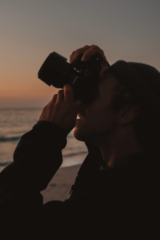 A person is holding a camera up to their eye, capturing a photo against the backdrop of a sunset over the ocean. The silhouette of the photographer is visible, adding a dramatic effect to the scene.