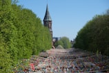 A large outdoor festival or event takes place under strings of colorful triangular flags. The scene is surrounded by lush green trees, and in the background, there is a tall church tower. The area is crowded with people, creating a vibrant and lively atmosphere.