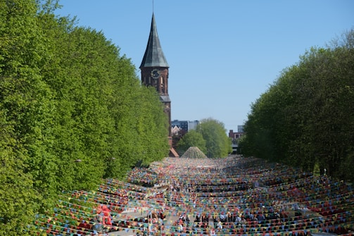 A large outdoor festival or event takes place under strings of colorful triangular flags. The scene is surrounded by lush green trees, and in the background, there is a tall church tower. The area is crowded with people, creating a vibrant and lively atmosphere.