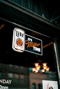 A neon sign hanging in a window displays the words 'Lite' and 'It's Miller Time' with a logo featuring a circular design. Behind the glass, a reflection of a building and interior lighting with a chandelier can be seen.