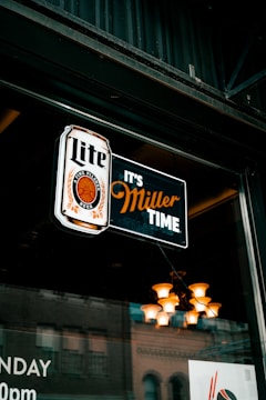 A neon sign hanging in a window displays the words 'Lite' and 'It's Miller Time' with a logo featuring a circular design. Behind the glass, a reflection of a building and interior lighting with a chandelier can be seen.