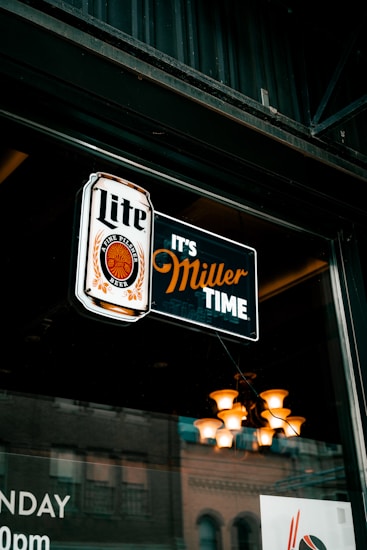 A neon sign hanging in a window displays the words 'Lite' and 'It's Miller Time' with a logo featuring a circular design. Behind the glass, a reflection of a building and interior lighting with a chandelier can be seen.