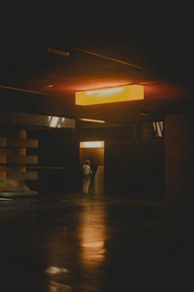 Dimly lit interior scene with a solitary figure standing at an ATM or kiosk. Overhead, a warm orange light casts a glow, creating soft reflections on the polished floor. The setting appears modern and minimalistic with geometric and textured architectural features.