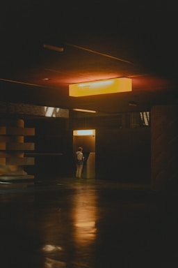 Dimly lit interior scene with a solitary figure standing at an ATM or kiosk. Overhead, a warm orange light casts a glow, creating soft reflections on the polished floor. The setting appears modern and minimalistic with geometric and textured architectural features.