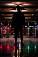 A silhouette of a person standing in front of illuminated turnstiles at a transit station, with another individual visible to the left. The setting is dimly lit with reflective, checkered flooring and various directional lights casting colorful glows.