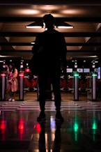 A silhouette of a person standing in front of illuminated turnstiles at a transit station, with another individual visible to the left. The setting is dimly lit with reflective, checkered flooring and various directional lights casting colorful glows.