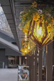 An artistic outdoor setting with decorative hanging lights. The lights are enclosed in wooden geometric frames and adorned with lush greenery and vibrant yellow flowers. This design creates a warm and inviting atmosphere along a wooden walkway, complemented by the soft glow of the bulbs.