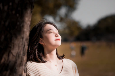 A peaceful woman sitting outside in nature, eyes closed in prayer with sunlight filtering through trees.