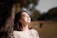 A smiling woman sitting outdoors with sunlight filtering through trees, looking refreshed and peaceful.