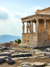 An ancient Greek architectural structure featuring a row of Caryatid columns stands prominently amidst scattered stone ruins. The backdrop includes a distant view of a hazy mountain range under a partly cloudy sky.