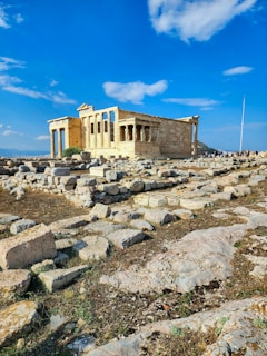 An ancient stone temple stands prominent under a bright, blue sky, surrounded by scattered rocks and ruins on the ground. The structure features classical architectural elements such as columns and sculptures.