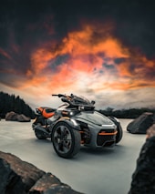 A futuristic three-wheeled vehicle is prominently displayed against a dramatic sky with vibrant orange and pink clouds. It is parked on a smooth surface with large rocks scattered around. The vehicle features sleek black and orange design elements, showcasing modern engineering.