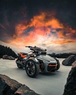 A futuristic three-wheeled vehicle is prominently displayed against a dramatic sky with vibrant orange and pink clouds. It is parked on a smooth surface with large rocks scattered around. The vehicle features sleek black and orange design elements, showcasing modern engineering.