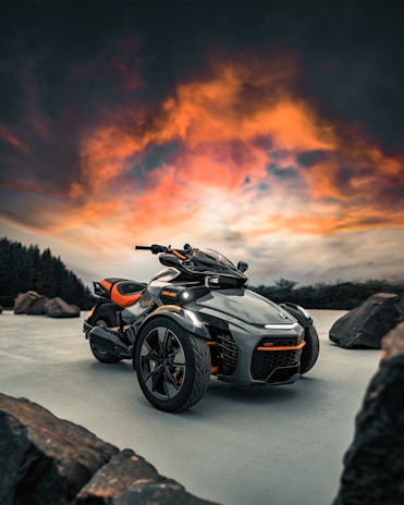 A futuristic three-wheeled vehicle is prominently displayed against a dramatic sky with vibrant orange and pink clouds. It is parked on a smooth surface with large rocks scattered around. The vehicle features sleek black and orange design elements, showcasing modern engineering.