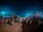 A nighttime market scene with people gathered around cattle. Wooden poles with tarps cover the stalls, illuminated by bright artificial lights against the dark sky. The ground is dirt, and children and adults are interacting with the cattle.