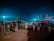 A nighttime market scene with people gathered around cattle. Wooden poles with tarps cover the stalls, illuminated by bright artificial lights against the dark sky. The ground is dirt, and children and adults are interacting with the cattle.