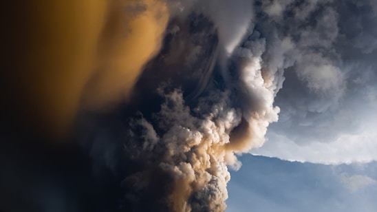 Volcanic eruption with dense plumes of dark and light ash against a backdrop of clear sky. The swirling clouds create a dramatic visual contrast and suggest intense natural activity.