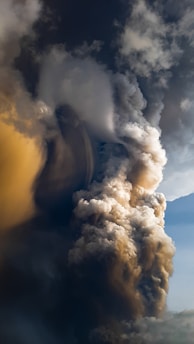 A towering, dense plume of smoke and ash rises dramatically into the sky, with swirling gray, white, and brown tones. The formation has a powerful and tumultuous appearance, conveying a sense of dynamic motion.