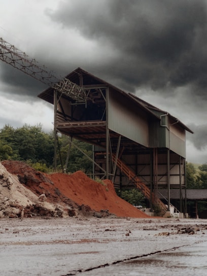 An industrial structure stands tall against a backdrop of dark, ominous clouds. The building is metal-framed with a complex network of beams and staircases. Piles of reddish-brown earth or sand are heaped in front, contrasting with the greenery of distant trees.