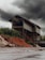 An industrial structure stands tall against a backdrop of dark, ominous clouds. The building is metal-framed with a complex network of beams and staircases. Piles of reddish-brown earth or sand are heaped in front, contrasting with the greenery of distant trees.