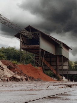 An industrial structure stands tall against a backdrop of dark, ominous clouds. The building is metal-framed with a complex network of beams and staircases. Piles of reddish-brown earth or sand are heaped in front, contrasting with the greenery of distant trees.
