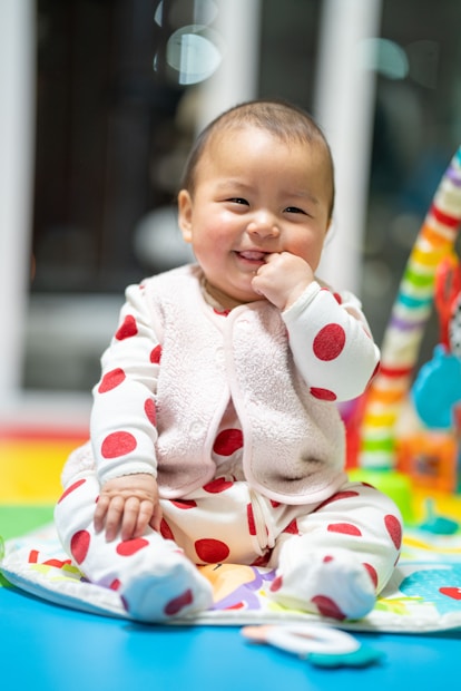 A smiling baby in a cozy outfit with red polka dots sits on a colorful play mat. The baby's expression is joyful and playful, with one hand near the mouth and a bright background filled with soft, blurred colors.