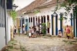 A cobblestone street flanked by colorful colonial buildings with vibrant blue and yellow doors. People are casually walking and socializing, with one person holding an umbrella. Plants and lanterns adorn the street, and a woman is sitting with a camera. The scene conveys a relaxed and historical ambiance.
